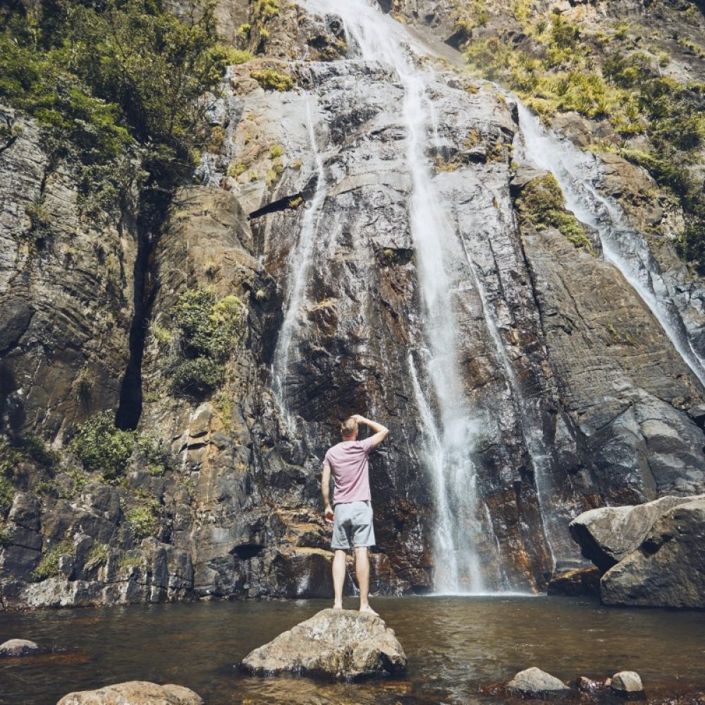 Young man (tourist) standing on stone in and watching stream of falling water in the middle of pure nature. Bambarakanda waterfalls - highest waterfall in Sri Lanka.